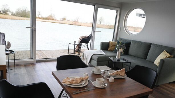 A dining area with a table set for two, a sofa, and a view of a body of water through large windows.