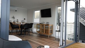 A modern living space with a dining area, a TV mounted on the wall, and a sliding glass door leading to an outdoor area.