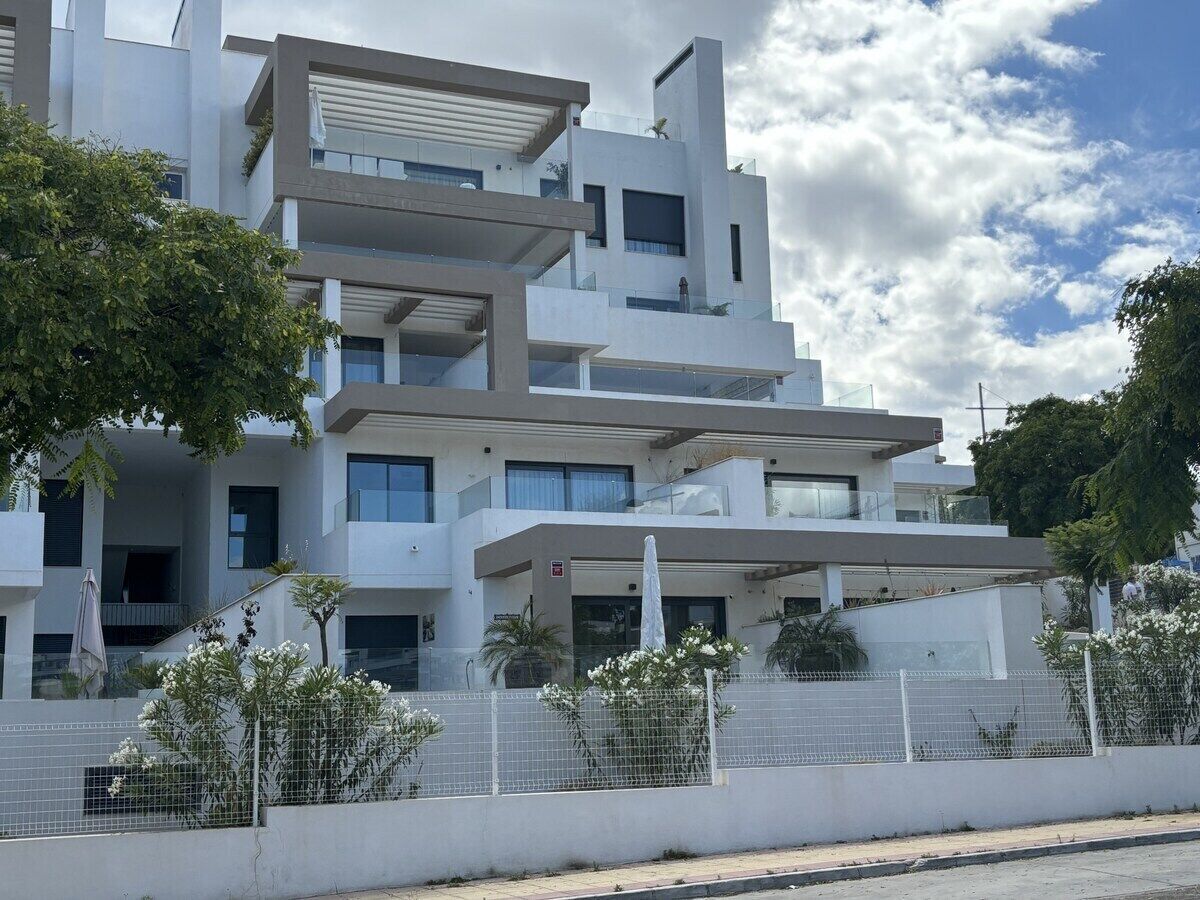 A modern multi-story residential building with balconies, a covered outdoor area, and a fenced garden.