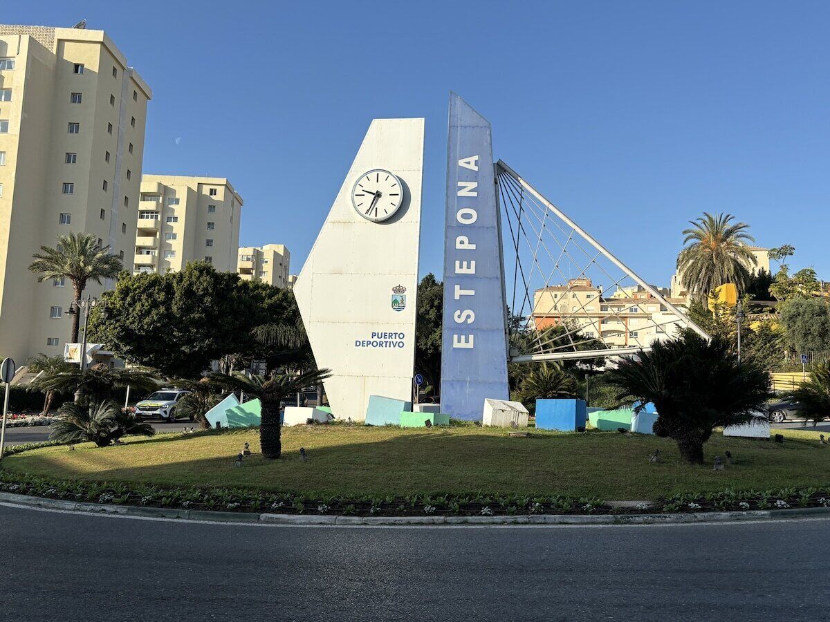 A sports monument with a clock, surrounded by greenery and buildings.