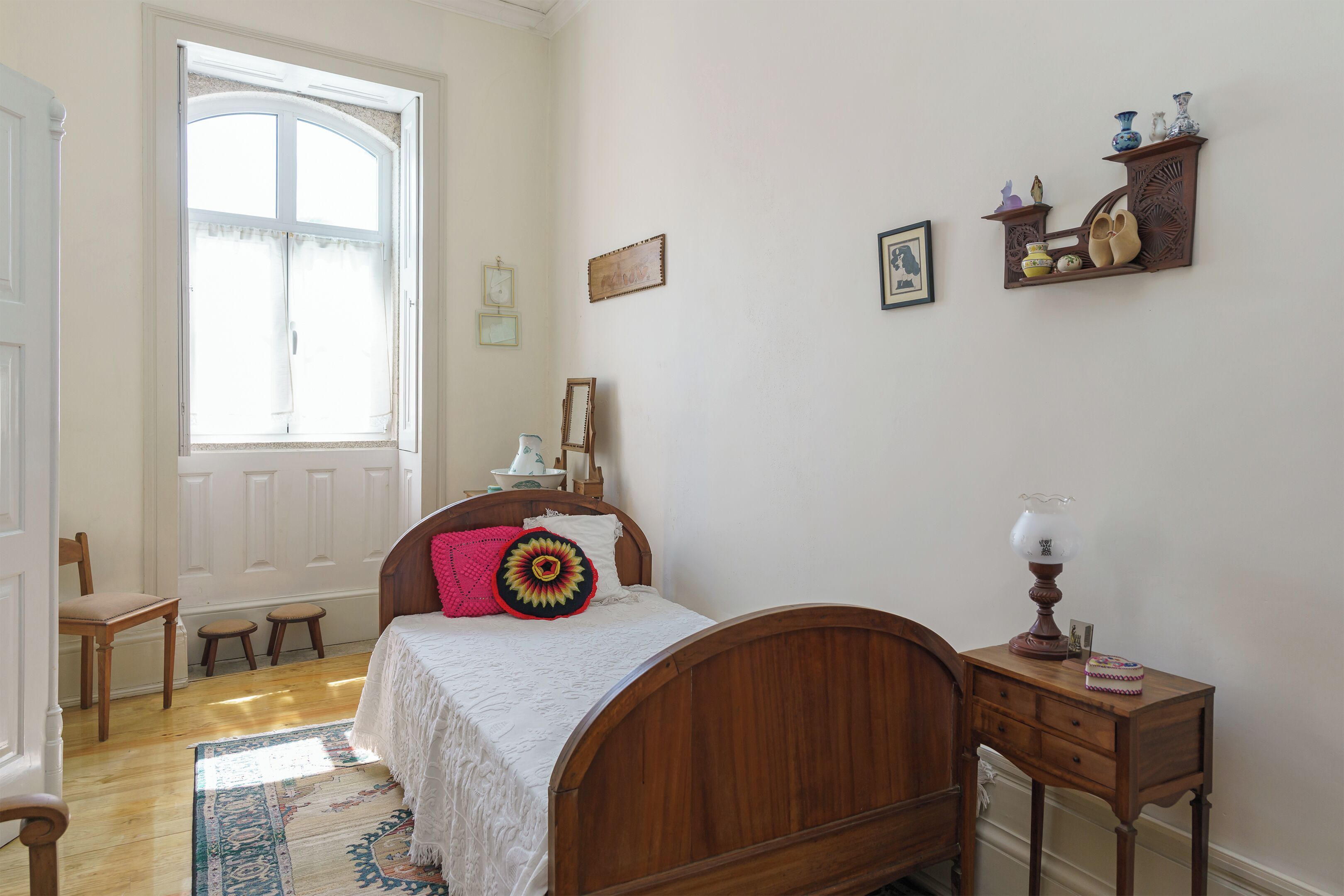 A bedroom with a wooden bed, a small bedside table, a nightstand, a window with curtains, and a door.