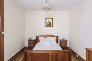 Een slaapkamer met een houten bed, witte beddengoed, een nachtkastje van hout met een klok, een houten dressoir en een kroonluchter.