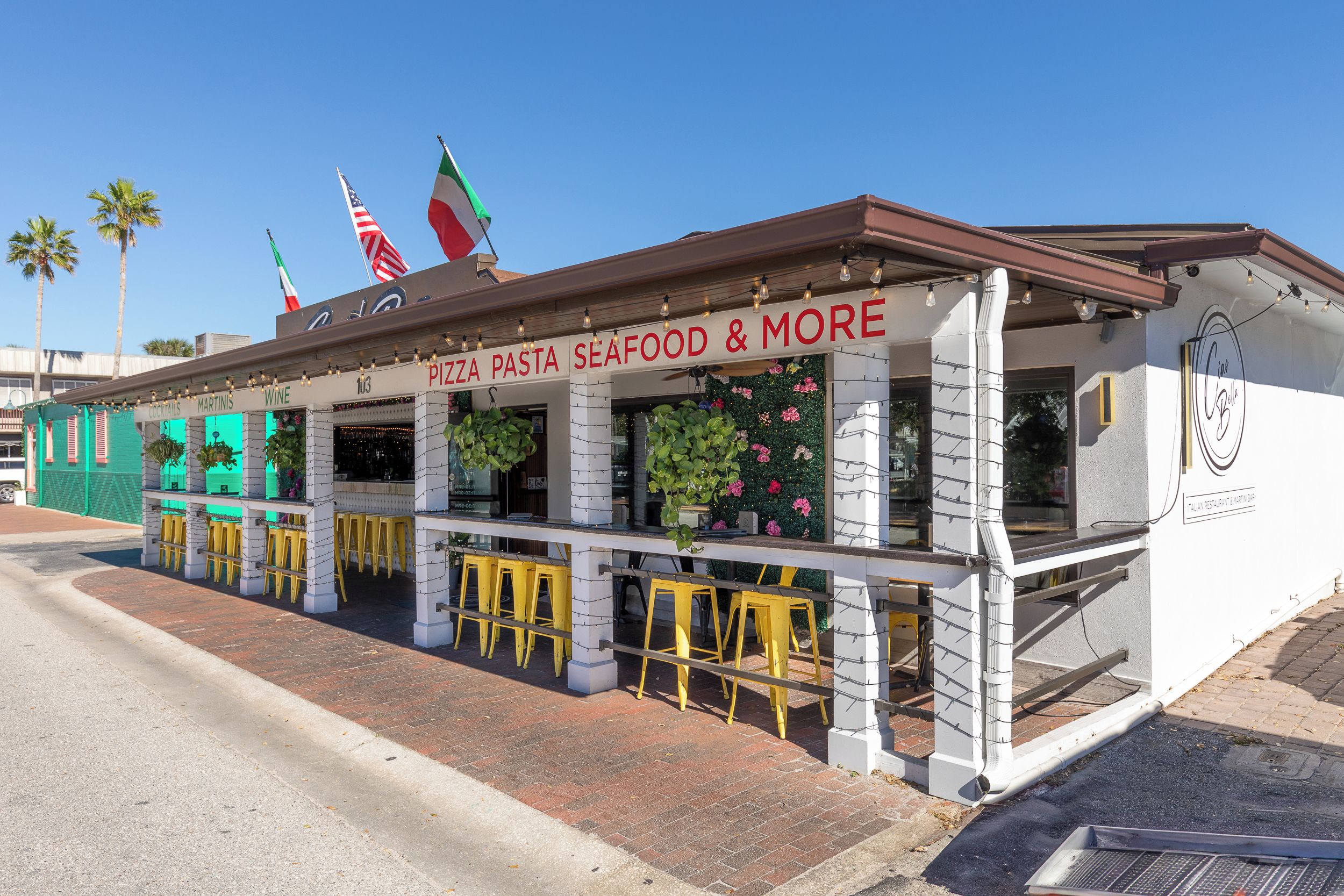 A seaside restaurant with outdoor seating and a sign reading 'PIZZA PASTA SEAFOOD & MORE'.