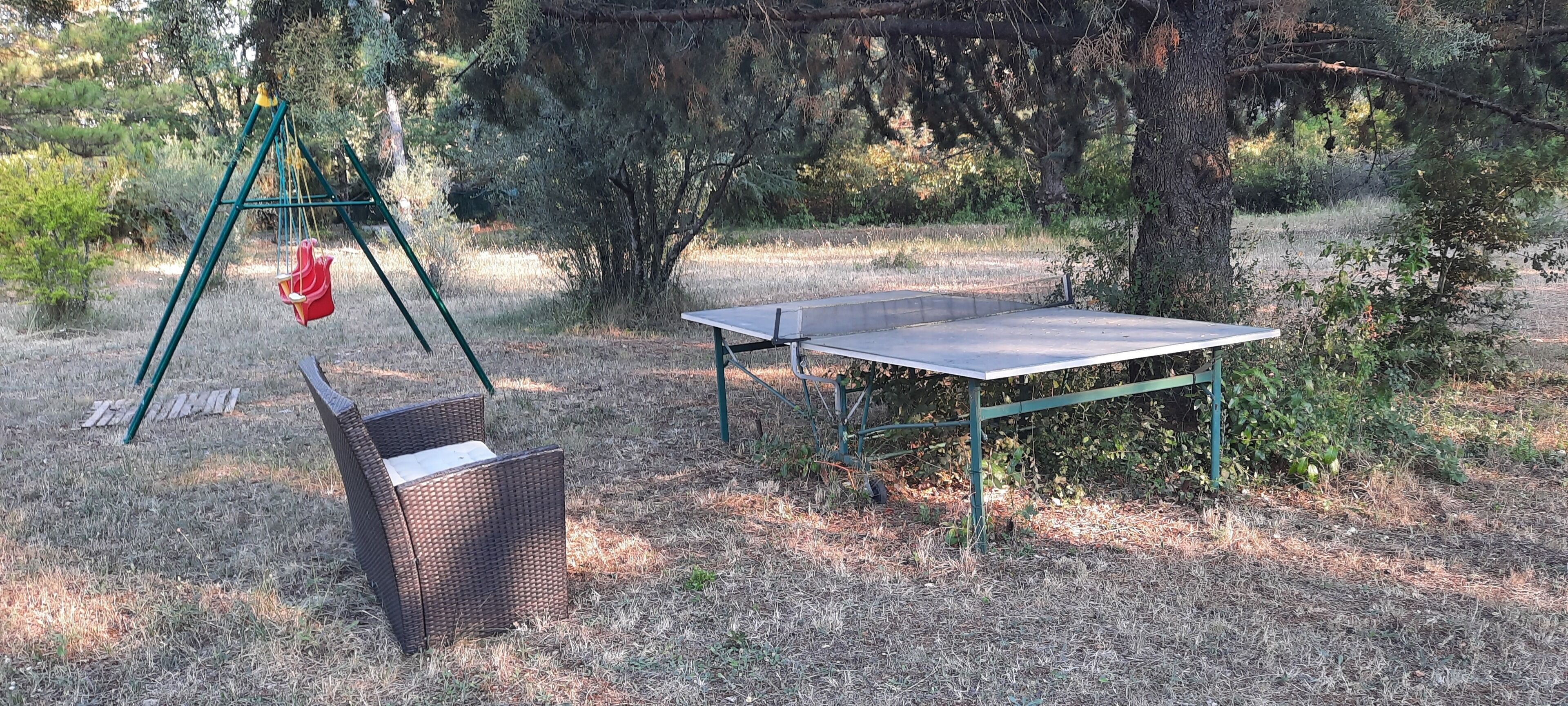A table tennis table with a green frame, a wicker chair, and a swing set in a wooded area.