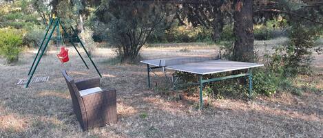 A table tennis table with a green frame, a wicker chair, and a swing set in a wooded area.