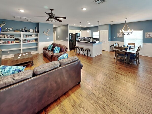 A spacious living area with a sofa, a coffee table, a bookshelf, a dining area with a table and chairs, and a kitchen with a bar. - Fin and Feather (Port Aransas)