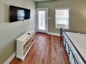 A bedroom with a bed, a TV mounted on the wall, a white dresser, and a window with blinds. - Fin and Feather (Port Aransas)