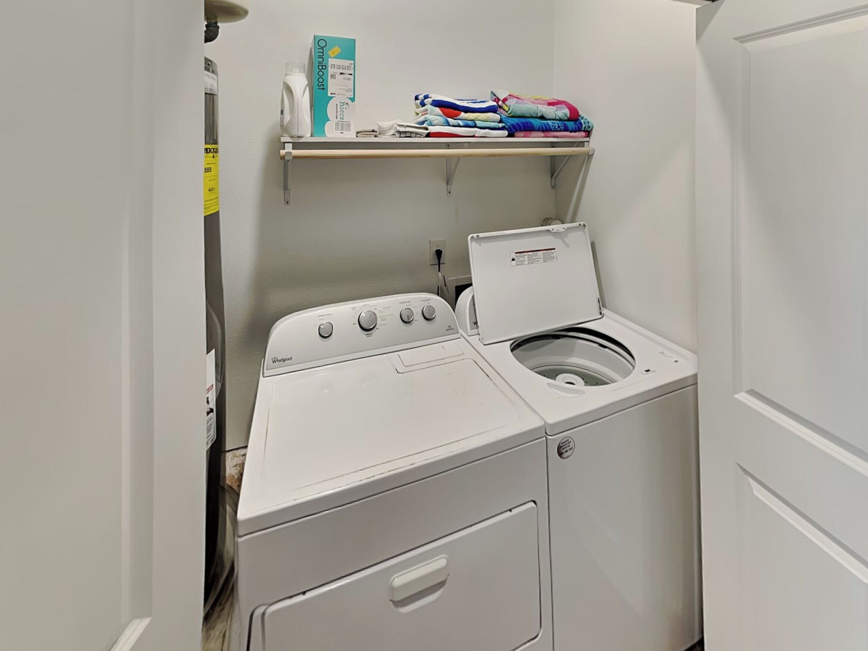 A compact laundry room with a front-loading washer and dryer, a shelf with folded clothes, and a box of laundry detergent.
