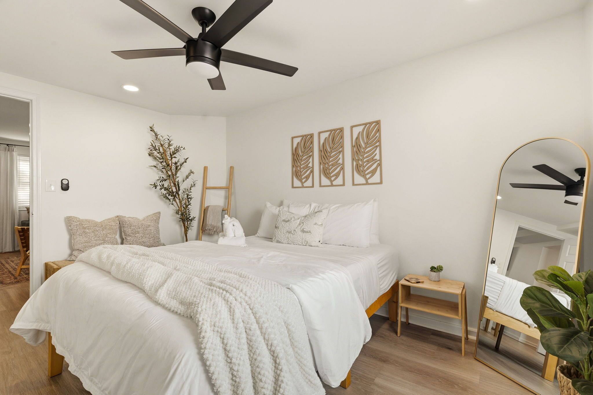 A bedroom with a bed, a ceiling fan, a bedside table, a mirror, and a plant.