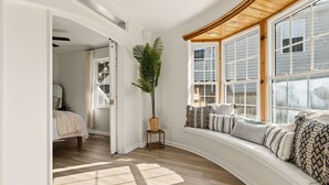 Una habitación con techo de madera curvada, ventanas amplias con contraventanas, una planta en maceta y un banco con cojines estampados.