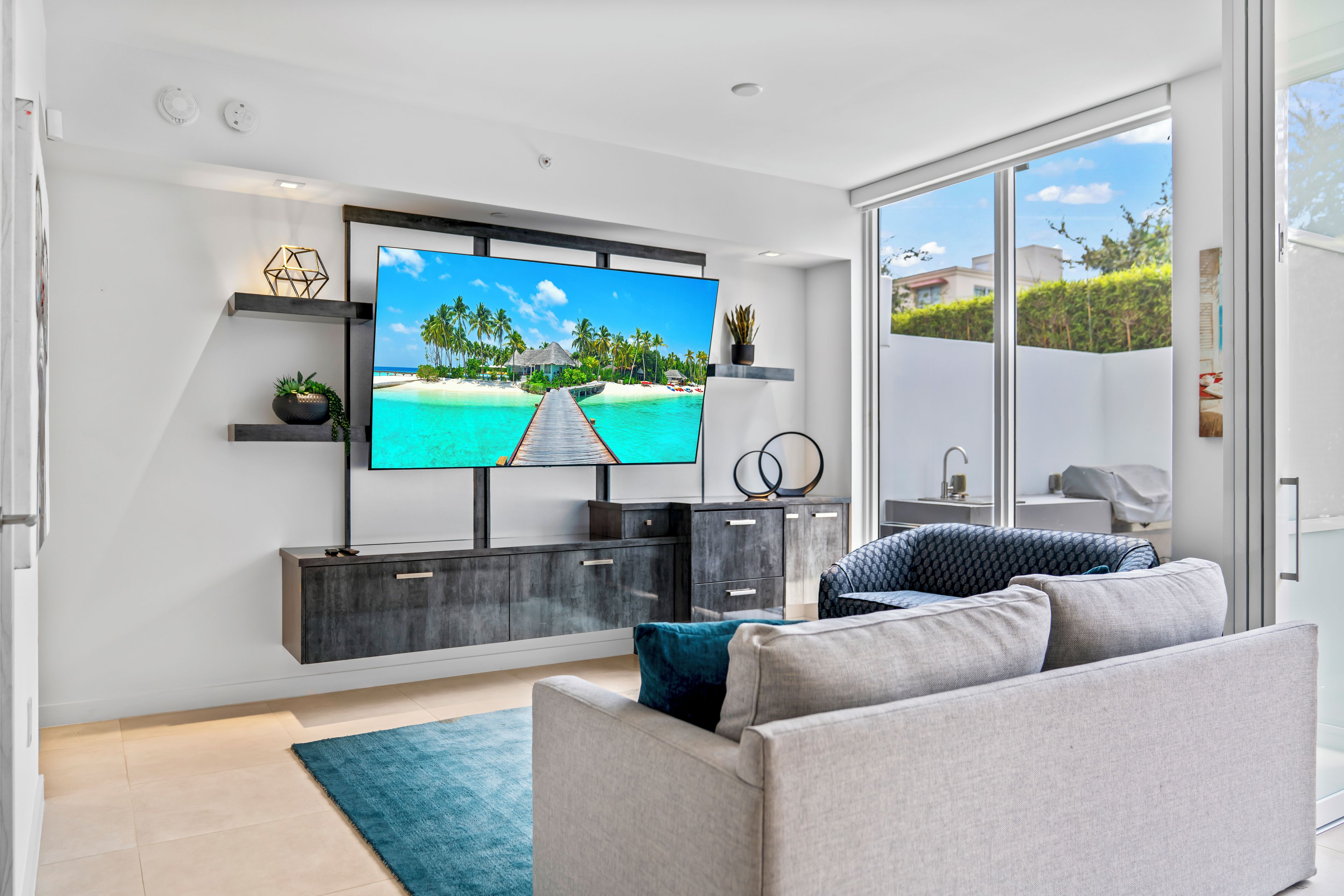 A modern living room with a flat-screen TV, grey sofa, and a view of a tropical beach through a sliding glass door.