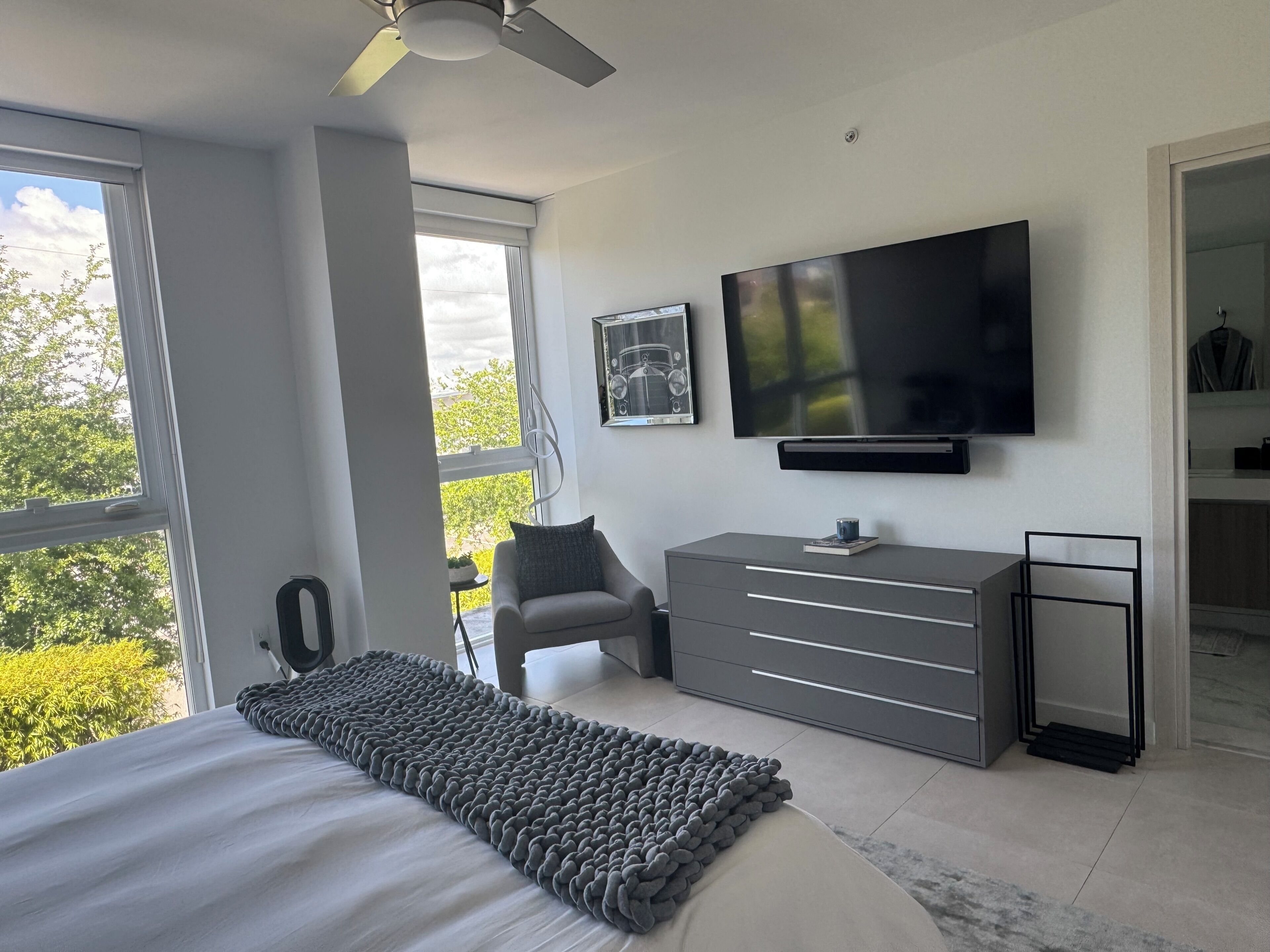 A modern bedroom with a large bed, a television, a dresser, and a view of greenery outside.
