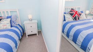 A bedroom with two beds, a nightstand with a lamp, and a mirror reflecting the room. - 2 Trelawny Way (Bembridge)