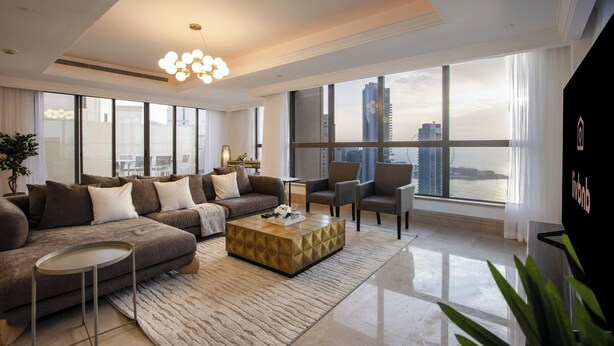 Ein modernes Wohnzimmer mit einer Sofagarnitur, Couchtisch und Sesseln, großen Fenstern mit Blick auf die Stadt und einer dekorativen Leuchte.