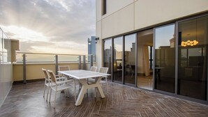 Ein moderner Balkon mit Holztable und -stühlen, einer Glastür, die zu einem Zimmer führt, und Blick auf die Stadtlandschaft.