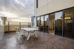 Ein moderner Balkon mit Holztable und -stühlen, einer Glastür, die zu einem Zimmer führt, und Blick auf die Stadtlandschaft.