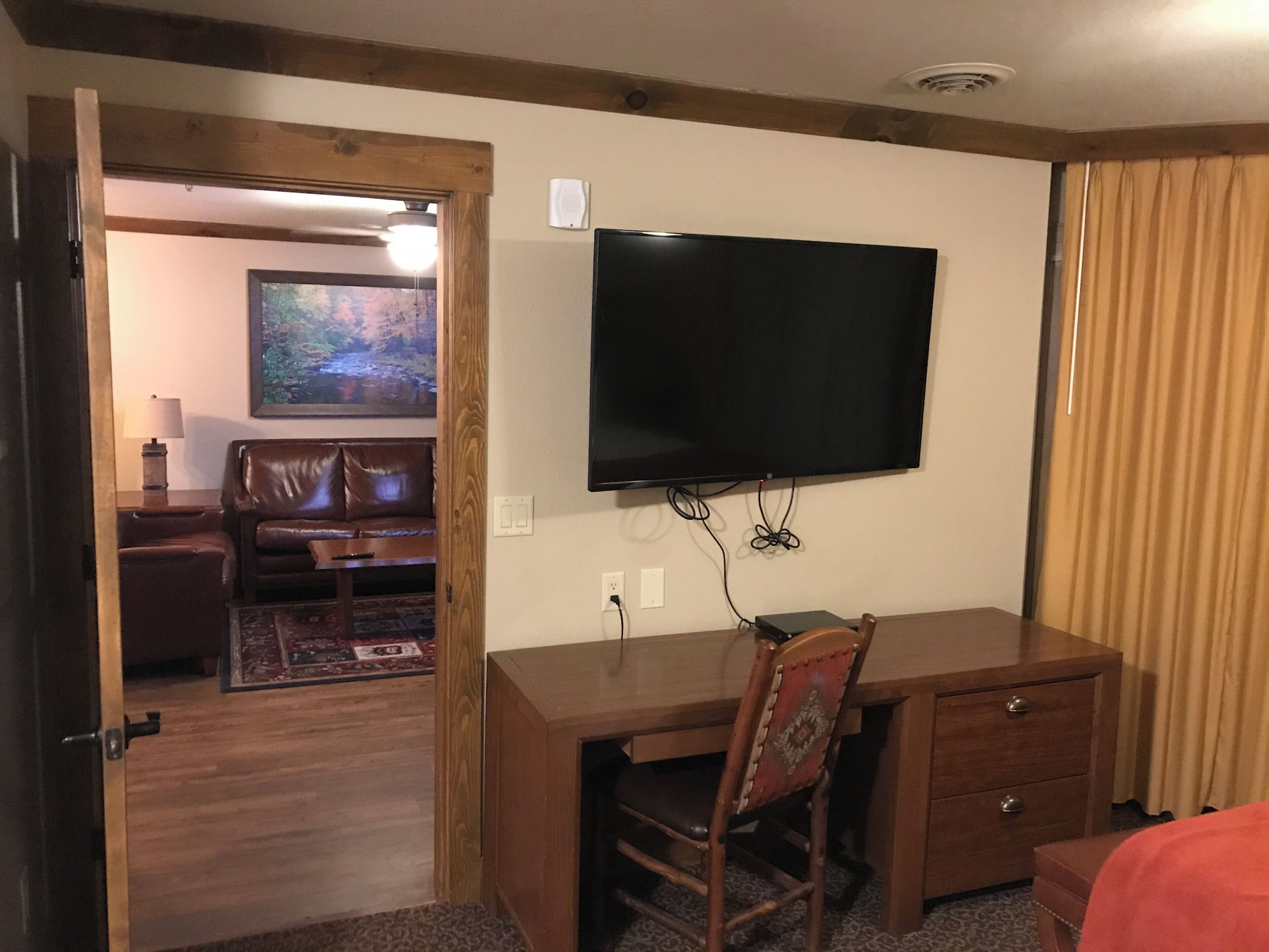Una habitación de hotel con un escritorio de madera, un televisor montado en la pared, un sillón de cuero, una mesita con una lámpara y un cuadro en la pared.