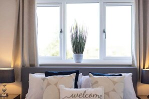 A room with a bed, a window with curtains, a potted plant, and a 'welcome' pillow.