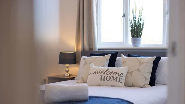 Una cama bien hecha con un cojín que dice 'bienvenido a casa', una lámpara de noche y una ventana con una planta.
