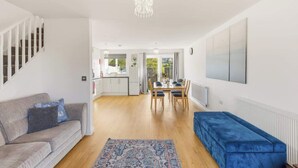 Una sala moderna con un sofá, un reposapiés azul, un tapete estampado, un comedor con una mesa y sillas, y una cocina al fondo.