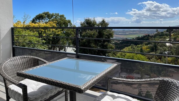 A balcony with wicker furniture, a glass table, and a view of a landscape with trees and buildings. - Sea and mountain view studio, new and comfortable (Saint-Paul)