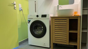 A compact laundry area with a front-loading washing machine, a wooden cabinet with a sink, and a wall-mounted mirror. - Sea and mountain view studio, new and comfortable (Saint-Paul)