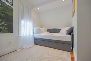 A bedroom with a bed, a bedside table, a window with curtains, and a wall-mounted light fixture.