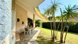 Un patio de piedra con mesa y sillas, una zona cubierta con techo blanco y un césped bien cuidado con palmeras.