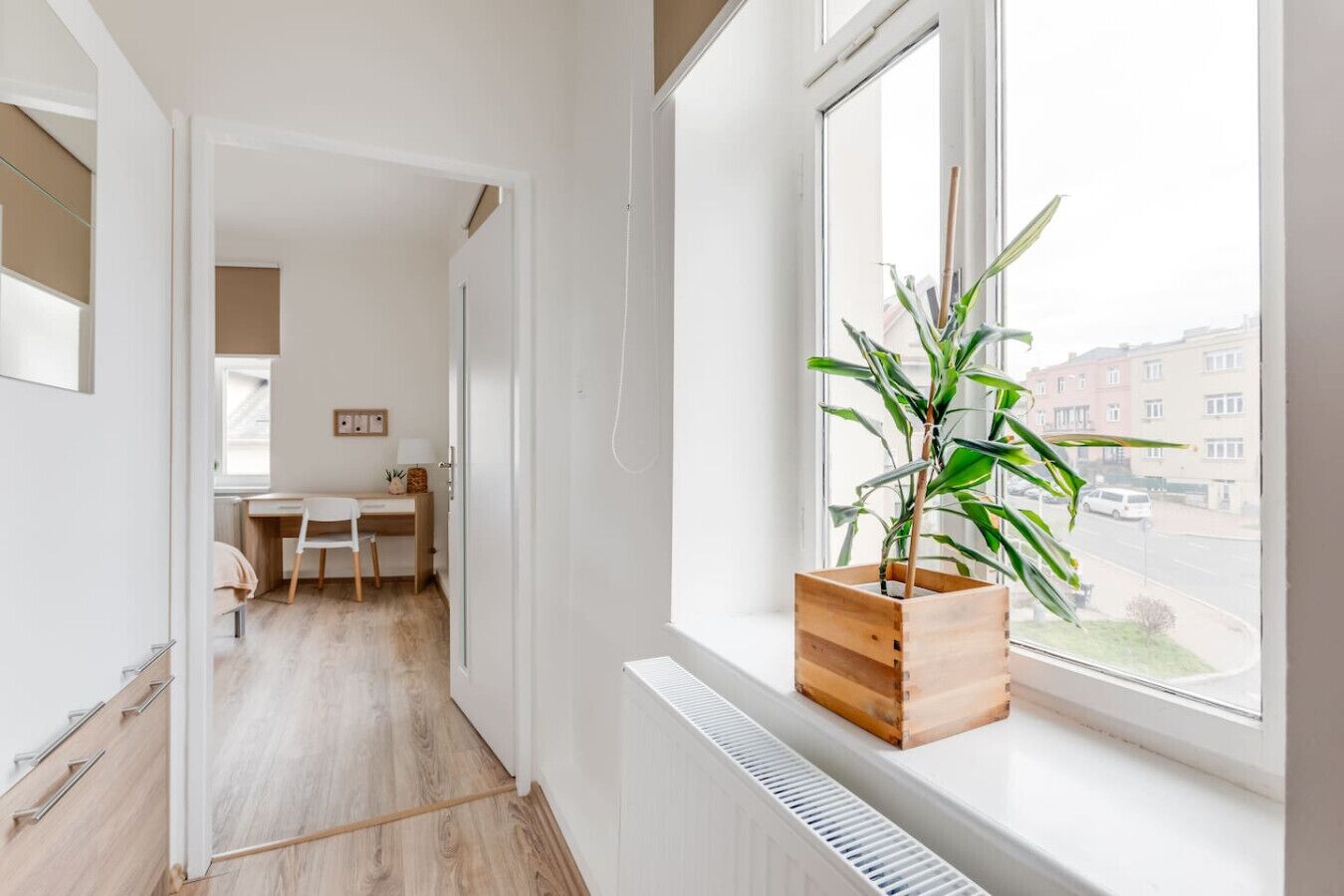 A modern apartment with a wooden floor, a desk with a chair, a radiator, and a window with a view of buildings and a street.