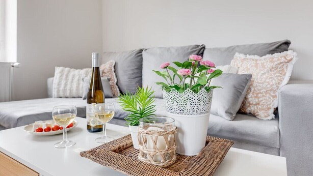 Moderni olohuone, jossa on harmaa sohva, valkoinen sohvapöytä, pullo valkoviiniä, kaksi lasia, lautanen kirsikkatomaatteja, ruukkukasvi ja koristeellinen maljakko.