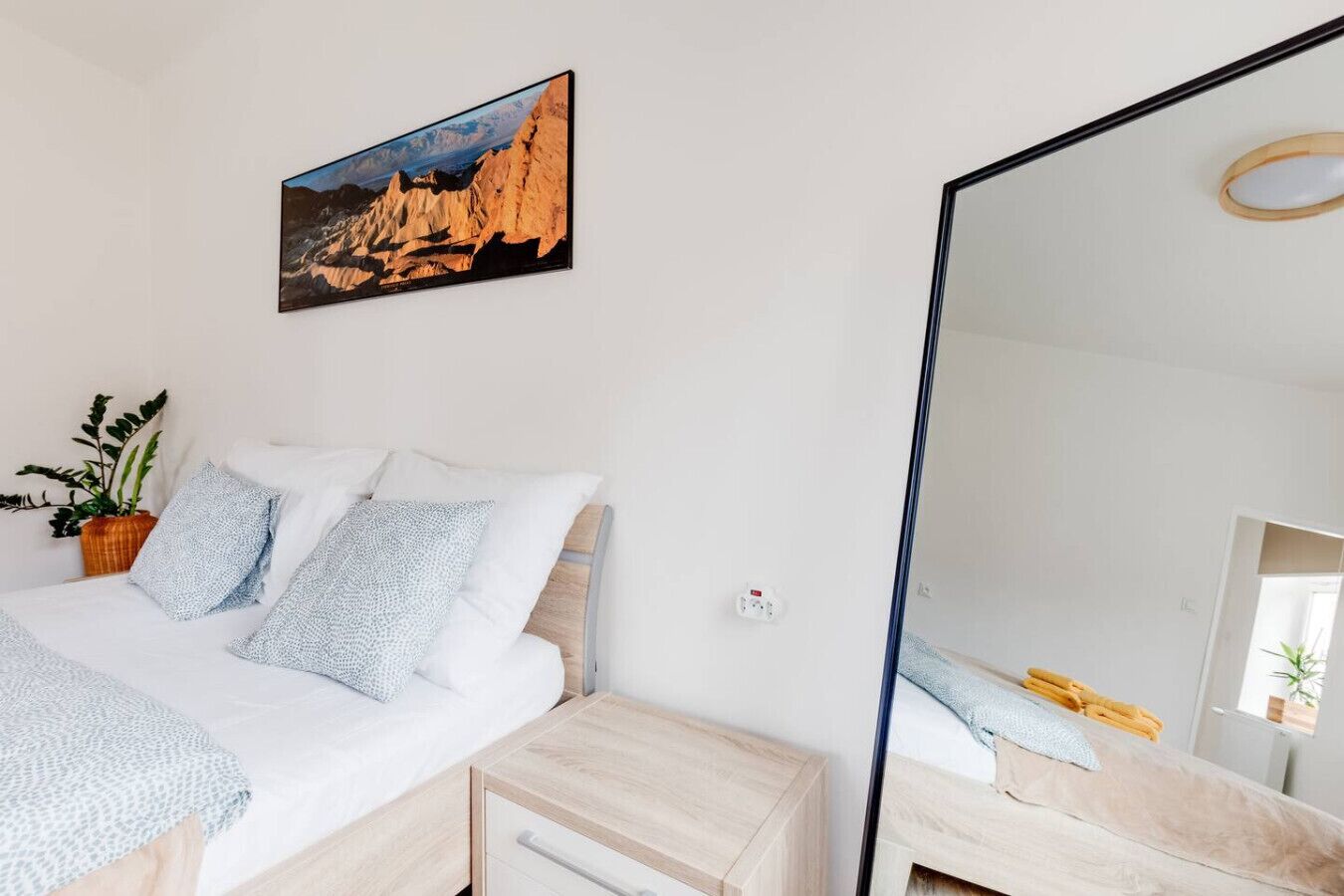 A bedroom with a bed, bedside tables, a mirror, a painting, and a potted plant.