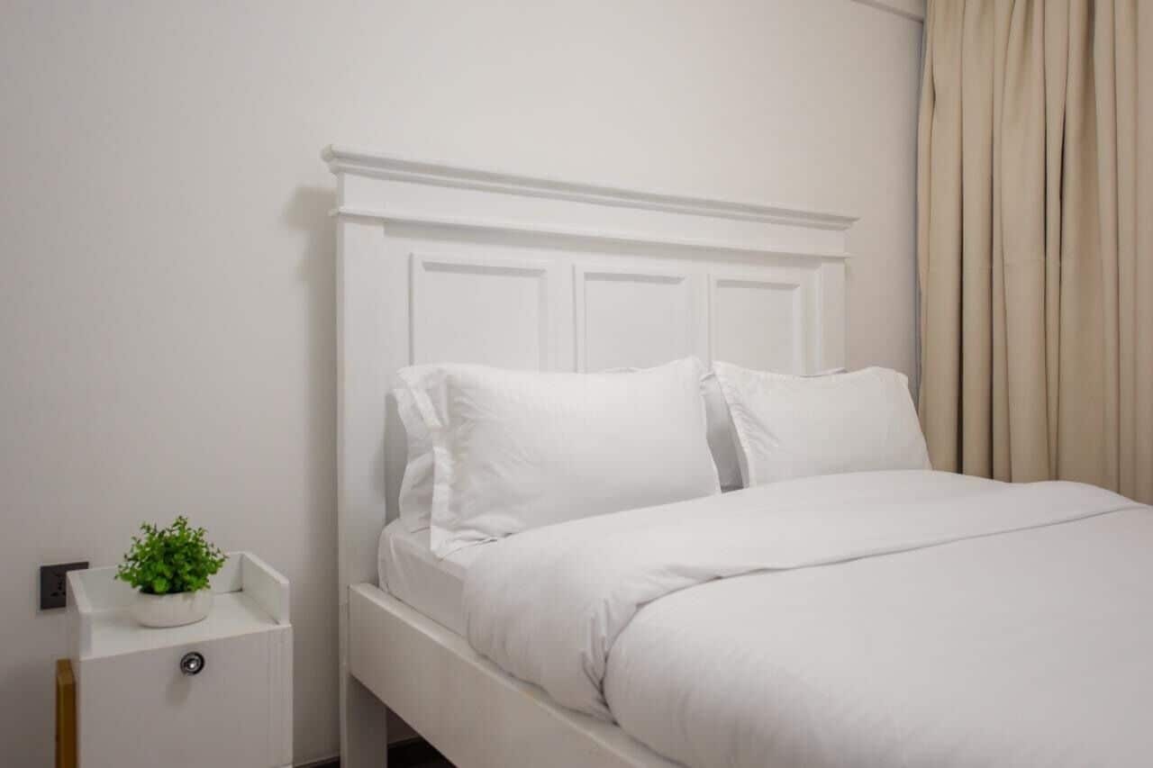 A white bed with a simple headboard, a bedside table with a plant, and a curtain on the window.