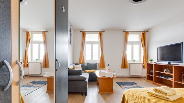 Moderni hotellihuone, jossa on sohva, sohvapöytä, televisio ja suuri ikkuna, jossa on verhot.