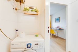 A laundry area with a washing machine, a toilet paper roll, and a shelf with plants labeled 'flowers'.