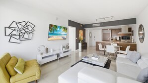 Una sala moderna con un sofá amarillo, sofás blancos y un comedor con sillas blancas. Se ve una cocina al fondo, con un microondas y armarios.