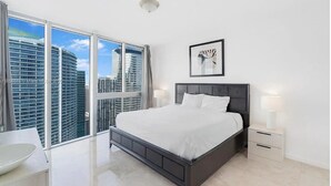 Un dormitorio moderno con una cama grande, mesitas de noche, una obra de arte enmarcada y vistas de rascacielos.