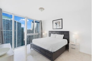 A modern bedroom with a large bed, bedside tables, a framed artwork, and a view of high-rise buildings.