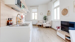 Une cuisine moderne dotée d’un four encastré, d’un coin repas avec une table et des chaises blanches, d’un téléviseur posé sur un meuble en bois et de grandes fenêtres.