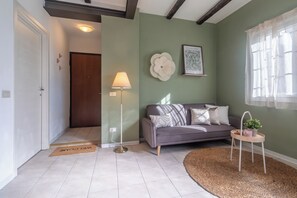 A living room with a sofa, a floor lamp, a welcome mat, a small table with a plant, and a framed botanical print on the wall.