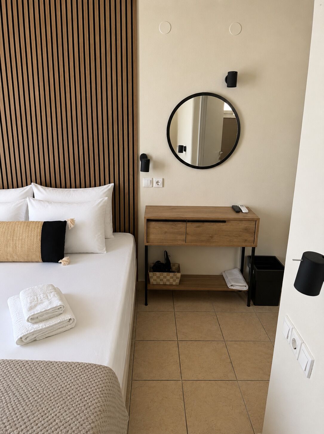 Une chambre d’hôtel comprenant un lit, une tête de lit en bois, un miroir rond, une coiffeuse en bois avec un lavabo et une poubelle.