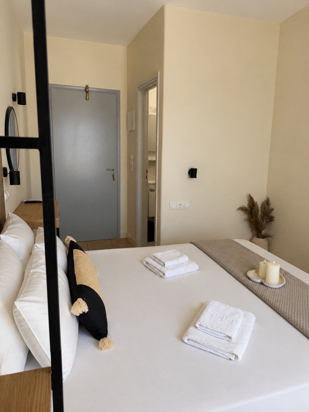 Quarto de hotel com cama, mesa de cabeceira, porta e luminária fixada na parede.