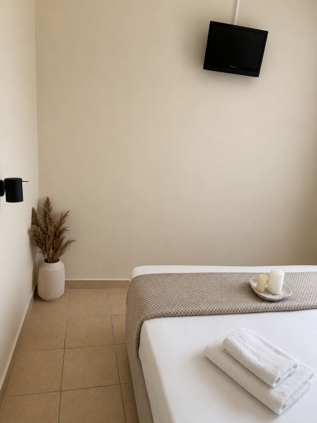 Une chambre d’hôtel au style minimaliste, comprenant un lit, un téléviseur à écran plat, un vase avec des plantes séchées et une petite table avec des bougies.