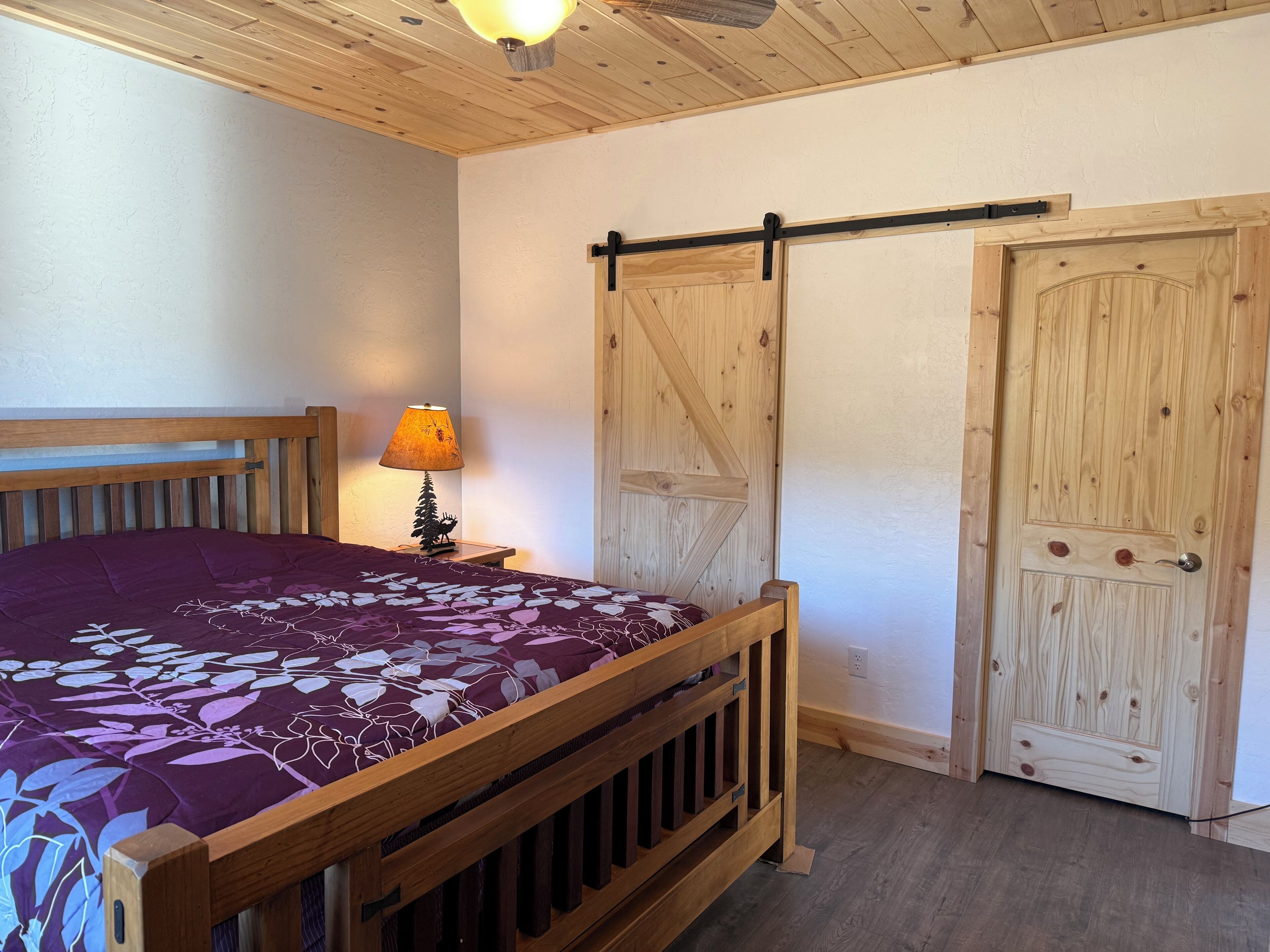Una cama de madera con colcha floral morada, una mesita de noche con lámpara, y una puerta de madera con puerta corredera de estilo rústico.