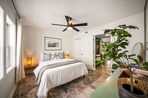 A bedroom with a bed, a ceiling fan, a bedside table with a lamp, a large potted plant, and a framed picture on the wall. - NEW! Driftwood Dunes - 3 Private Units - Sleeps 14 (Carolina Beach)