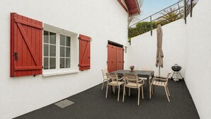 En uteplats med ett bord och stolar, en grill och röda fönsterluckor.