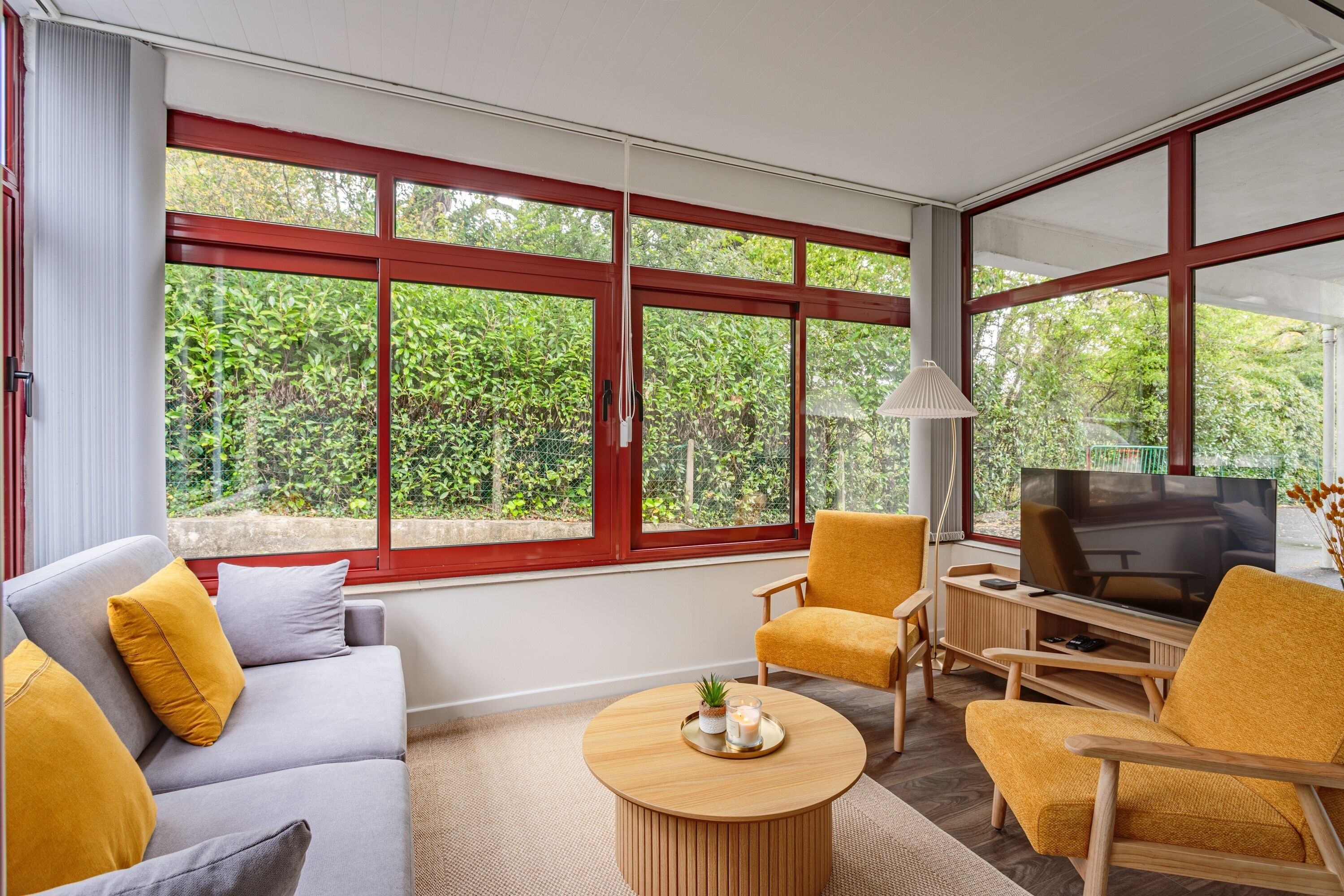 A modern living room with a sofa, armchairs, a coffee table, and a TV. Large windows offer a view of greenery outside.