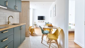 Een moderne keuken met grijze kastjes, een houten aanrecht en een spoelbak. Er is een eethoek met een ronde tafel en gele stoelen. Een televisie is aan de muur bevestigd op de achtergrond.