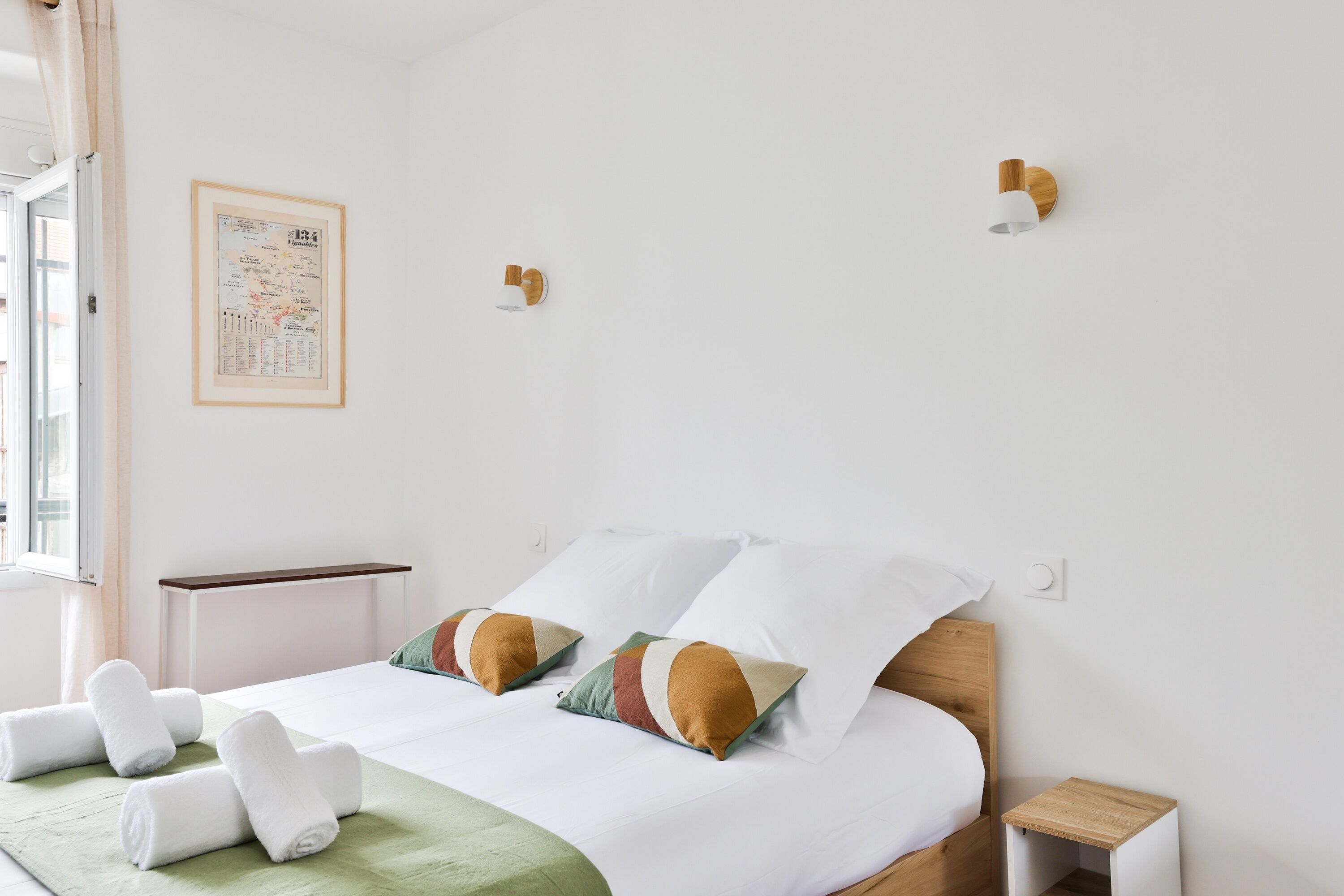 Un lit bien fait, avec du linge de lit blanc et deux coussins décoratifs, une petite table de chevet, un luminaire fixé au mur et une carte encadrée accrochée au mur.