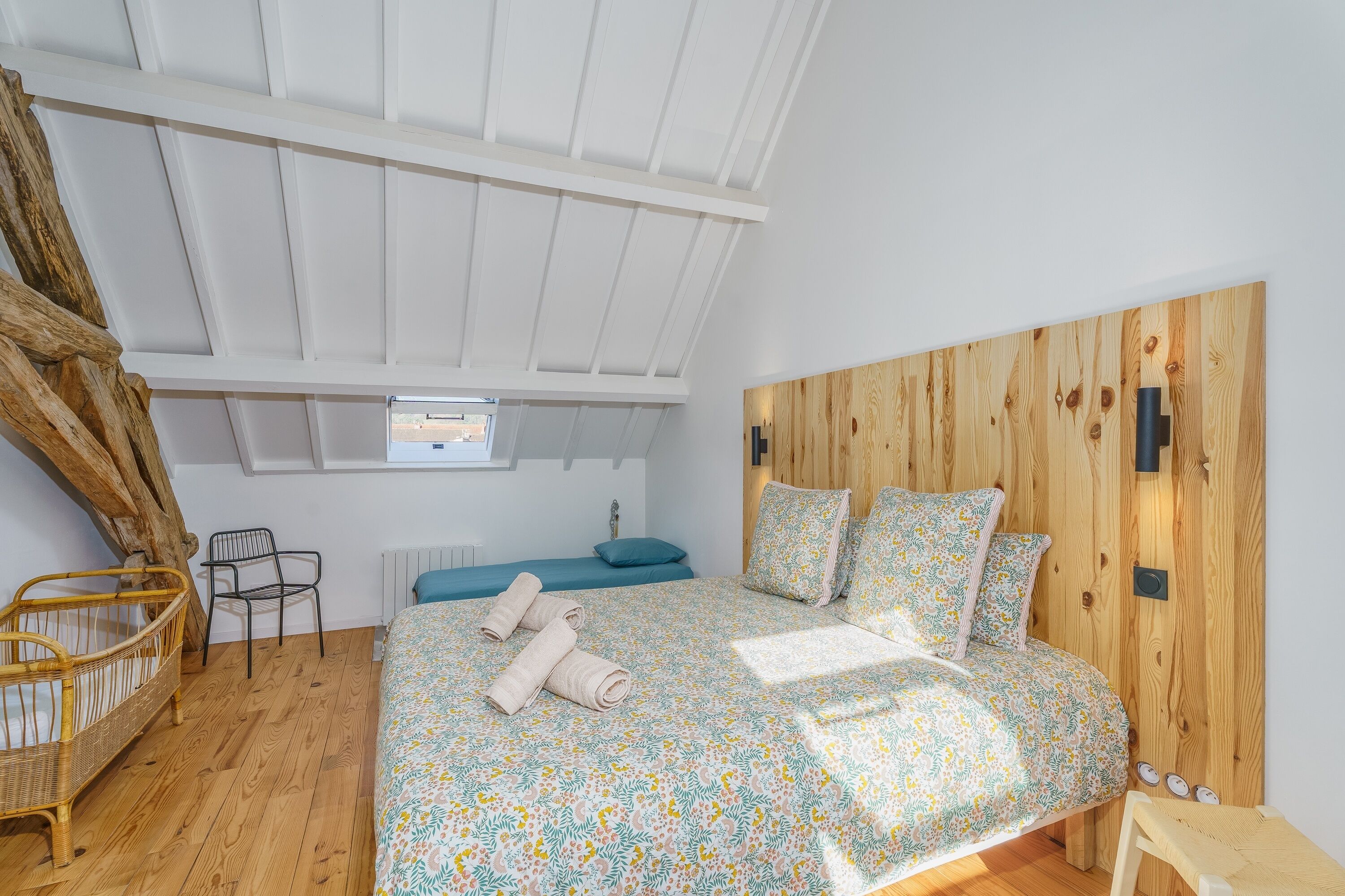 A bedroom with a wooden headboard, a bed with floral bedding, a crib, and a chair.
