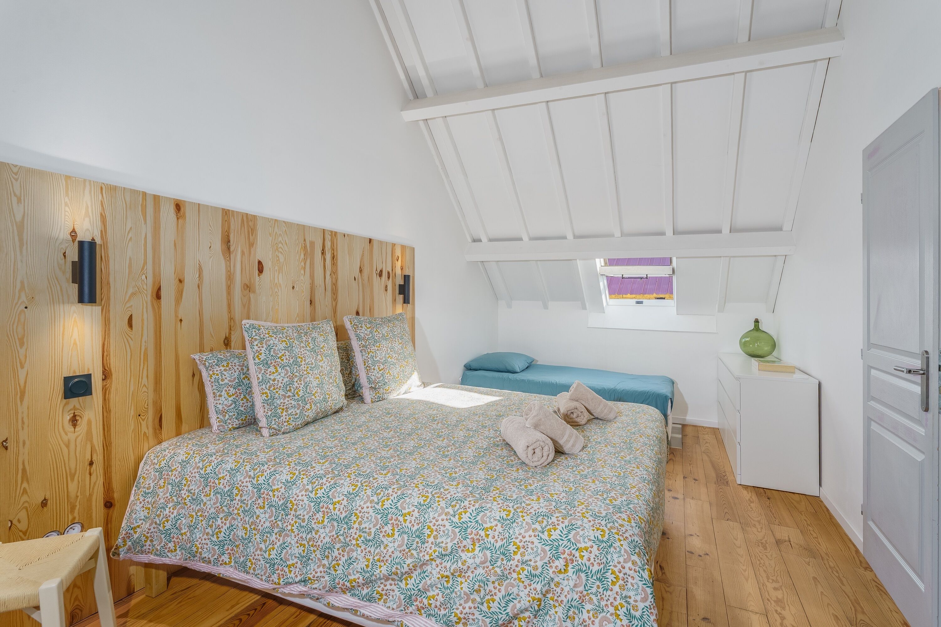 A bedroom with a wooden headboard, a bed with floral bedding, a small desk, and a window with a view.